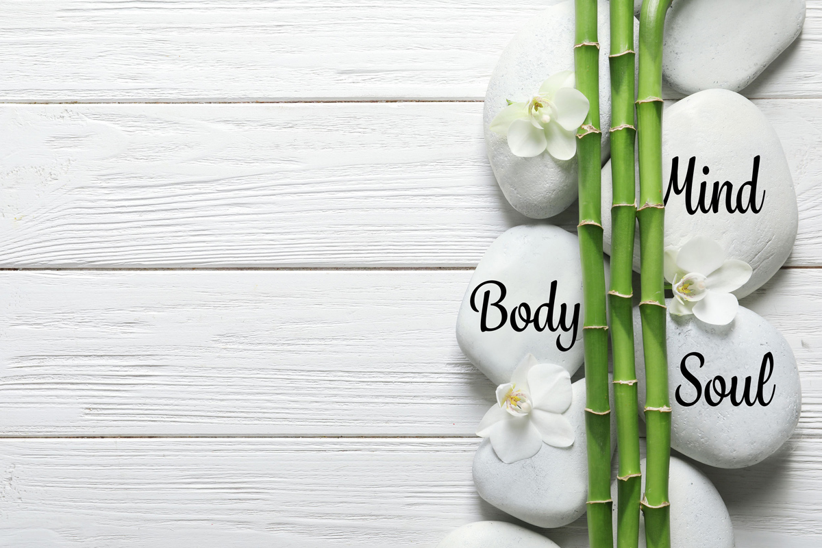White rocks that say “Mind, Body, Soul” with white flowers and bamboo laying on white boards.